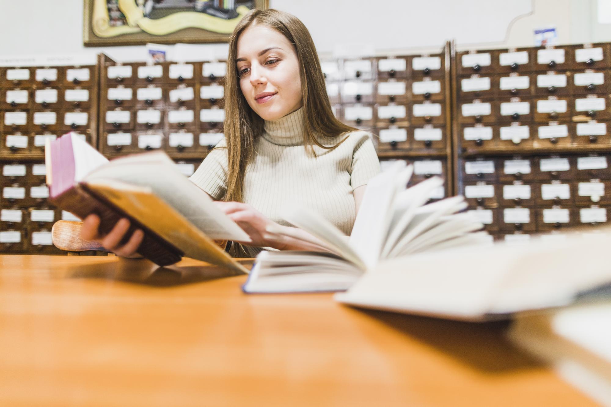 Kobieta siedząca przy biurku w bibliotece czyta książkę, dwie otwarte książki leżą na biurku, w tle katalog kartkowy.