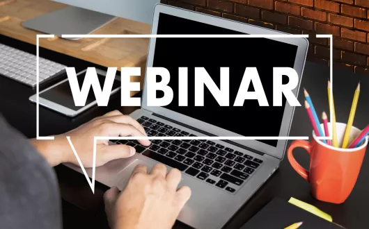 Napis „WEBINAR”, w tle laptop, kubek z długopisami, dłonie na klawiaturze.