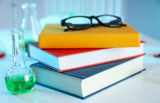 Zdjęcie poglądowe przedstawiające ułożone jedna na drugiej trzy książki, na wierzchu leżą okulary, obok książek stoi kolba z zielonym płynem.